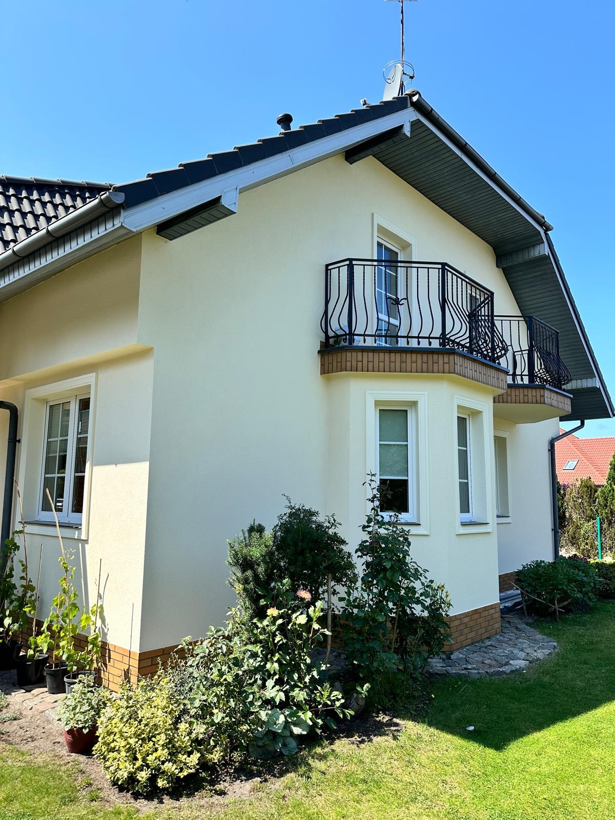 Elewacja jasnego domu jednorodzinnego z dwoma balkonami z ozdobnymi, czarnymi balustradami, widok z poziomu ogrodu z bujną roślinnością.