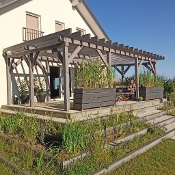 Zakład Stolarski Antoni Bojka - Nowoczesna pergola z szarego drewna, zintegrowana z tarasem domu. Rośliny w donicach na tarasie. Osoba siedząca przy stole. Błękitne niebo w tle.