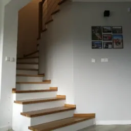 Nowoczesne schody z drewnianymi stopniami i białymi podstopnicami, z minimalistyczną balustradą z metalowych linek. Na ścianie galeria zdjęć w czarnych ramkach.