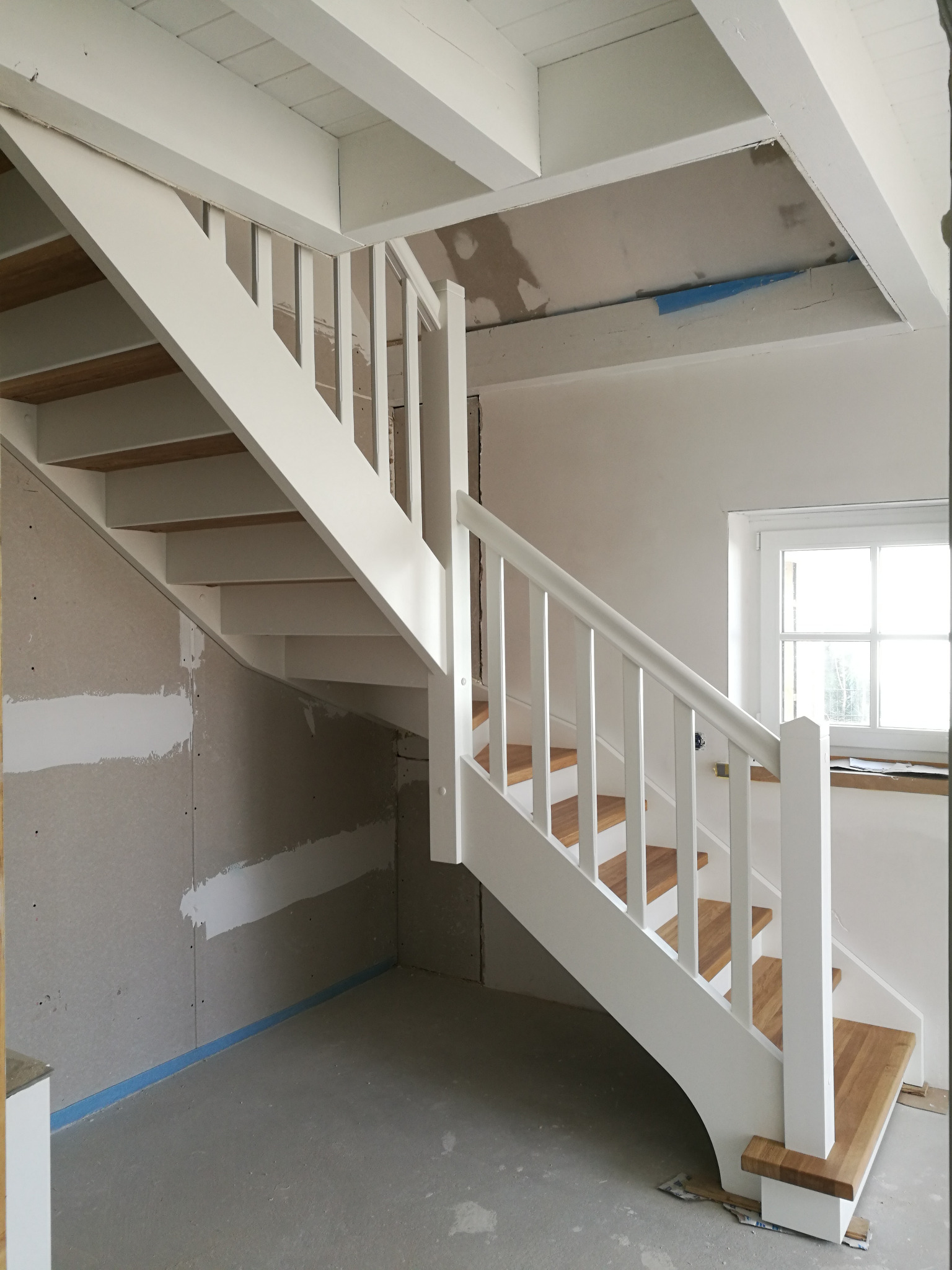 Białe schody z drewnianymi stopniami w surowym wnętrzu. Widoczne niedokończone ściany i sufit, podkreślające etap montażu. Minimalistyczny design, jasne oświetlenie.