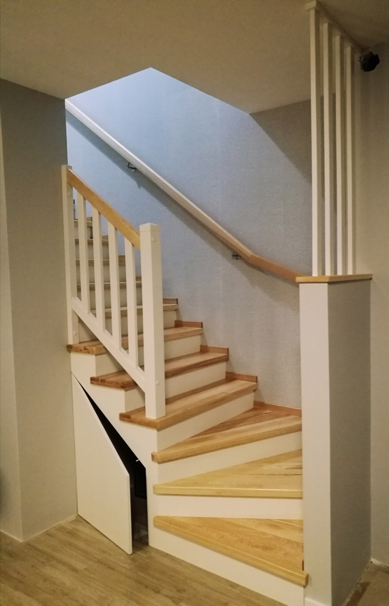 Jasne drewniane schody z białą balustradą i zabudową pod schodami z otwartymi drzwiczkami, widok z dołu. Neutralne tło ściany i podłogi.