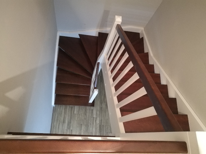 Widok z góry na kręte schody drewniane z białą balustradą. Ciemne stopnie kontrastują z jasnymi ścianami. Podłoga wykończona panelami imitującymi drewno.