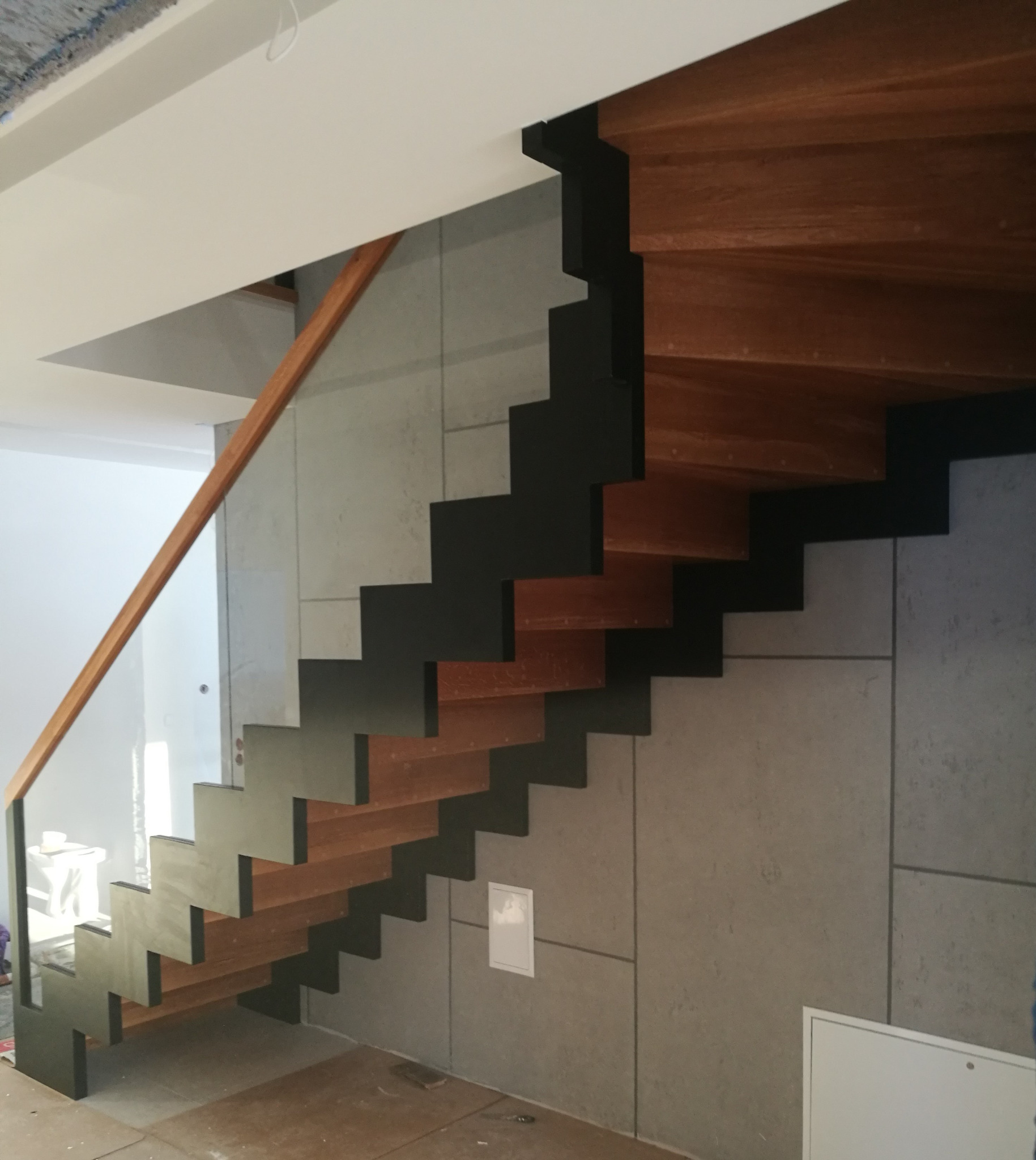 Nowoczesne schody wiszące z drewnianymi stopniami i czarną konstrukcją, zamontowane przy szarej ścianie. Minimalistyczny design, widoczna balustrada.