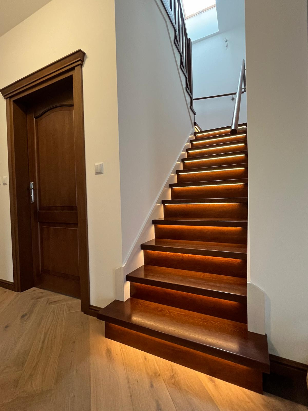 Oświetlone, ciemne schody z drewna prowadzą do góry. Widoczna balustrada i fragment drzwi z drewnianą ościeżnicą. Podłoga z parkietu ułożonego w jodełkę.