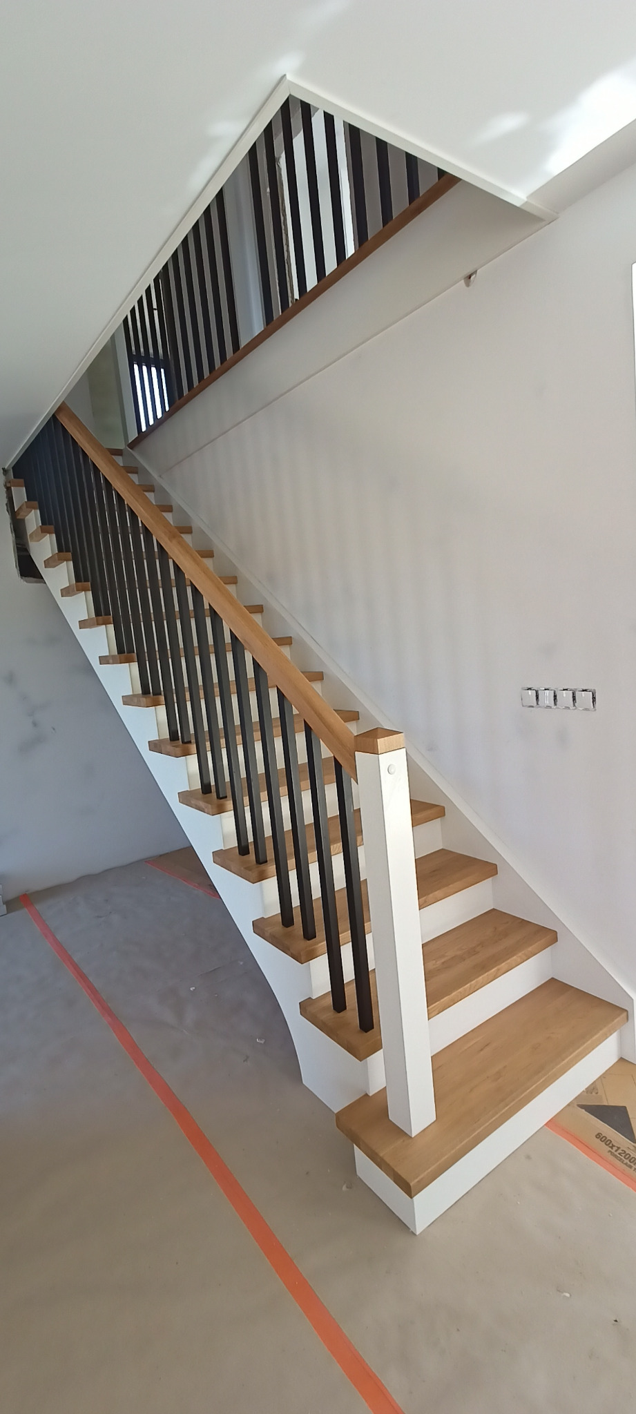 Nowoczesne schody z drewnianymi stopniami i czarną, metalową balustradą w jasnym wnętrzu. Widoczne zabezpieczenia podłogi podczas montażu.