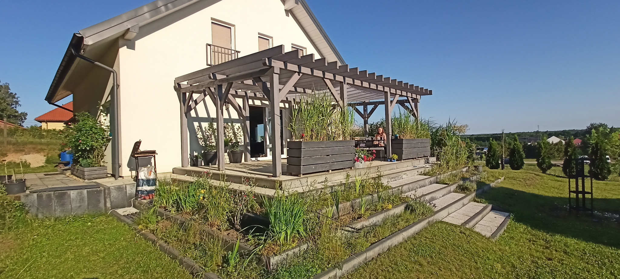 Nowoczesna pergola z szarego drewna, zintegrowana z tarasem domu. Rośliny w donicach na tarasie. Osoba siedząca przy stole. Błękitne niebo w tle.