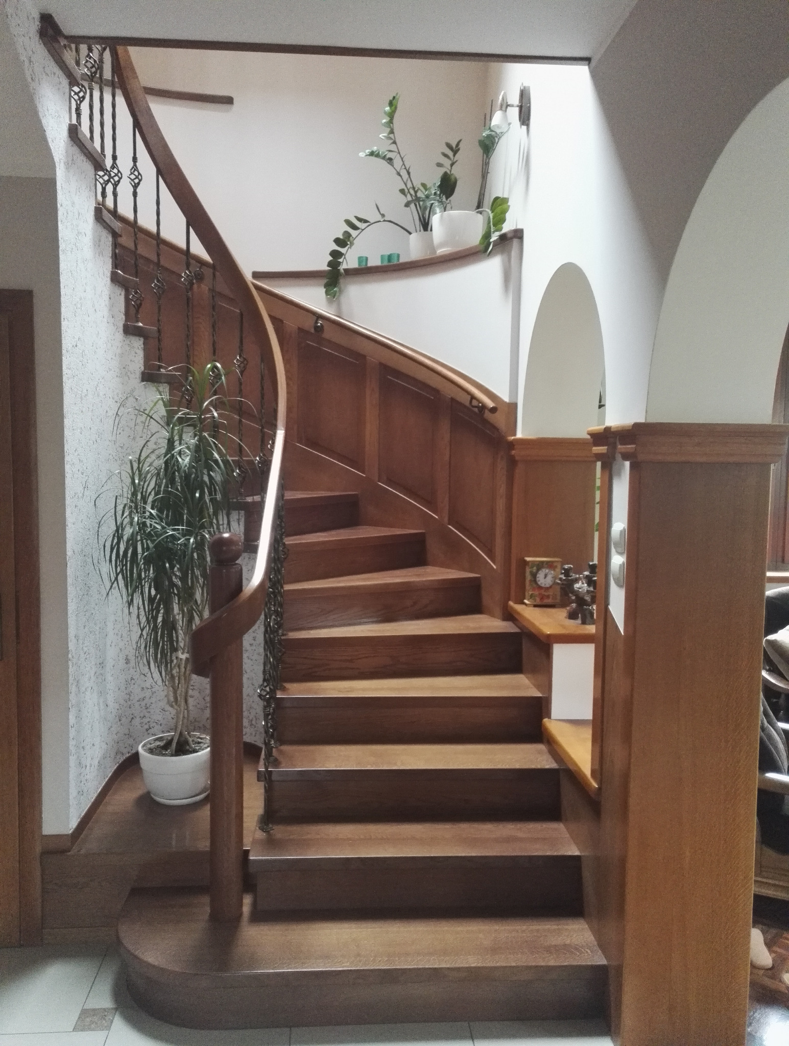 Kręte schody z drewna z kutą balustradą, oświetlone światłem wpadającym przez okno. Na podeście roślina doniczkowa. U dołu schodów kolejna roślina w doniczce.