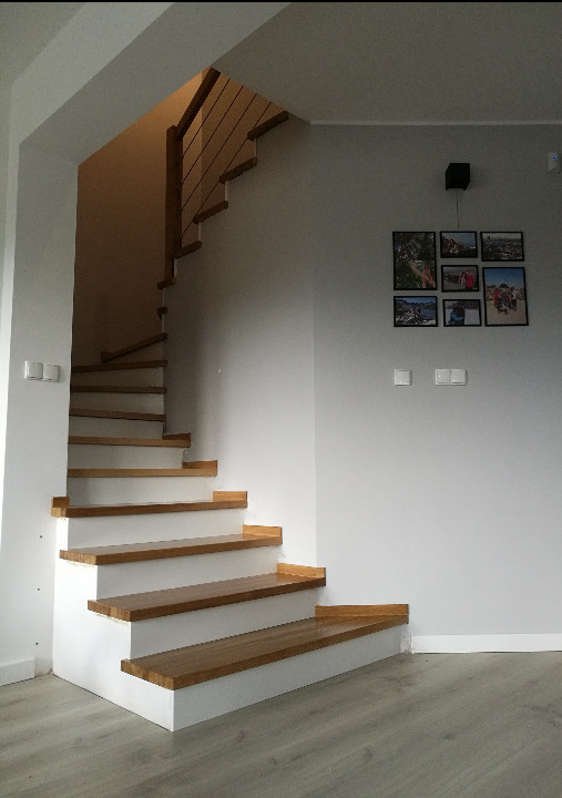 Nowoczesne schody z drewnianymi stopniami i białymi podstopnicami, z minimalistyczną balustradą z metalowych linek. Na ścianie galeria zdjęć w czarnych ramkach.