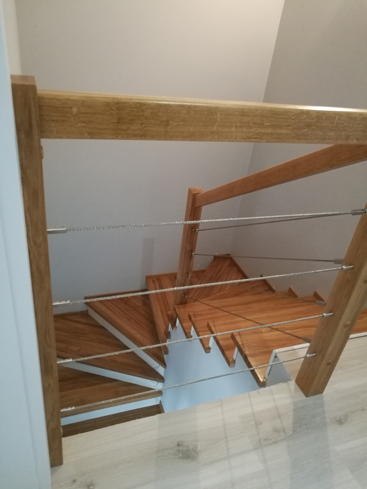 Kątowe ujęcie schodów z jasnego drewna z balustradą z drewnianych słupków i stalowych linek, patrząc z góry. Widoczny fragment podłogi i ściany.