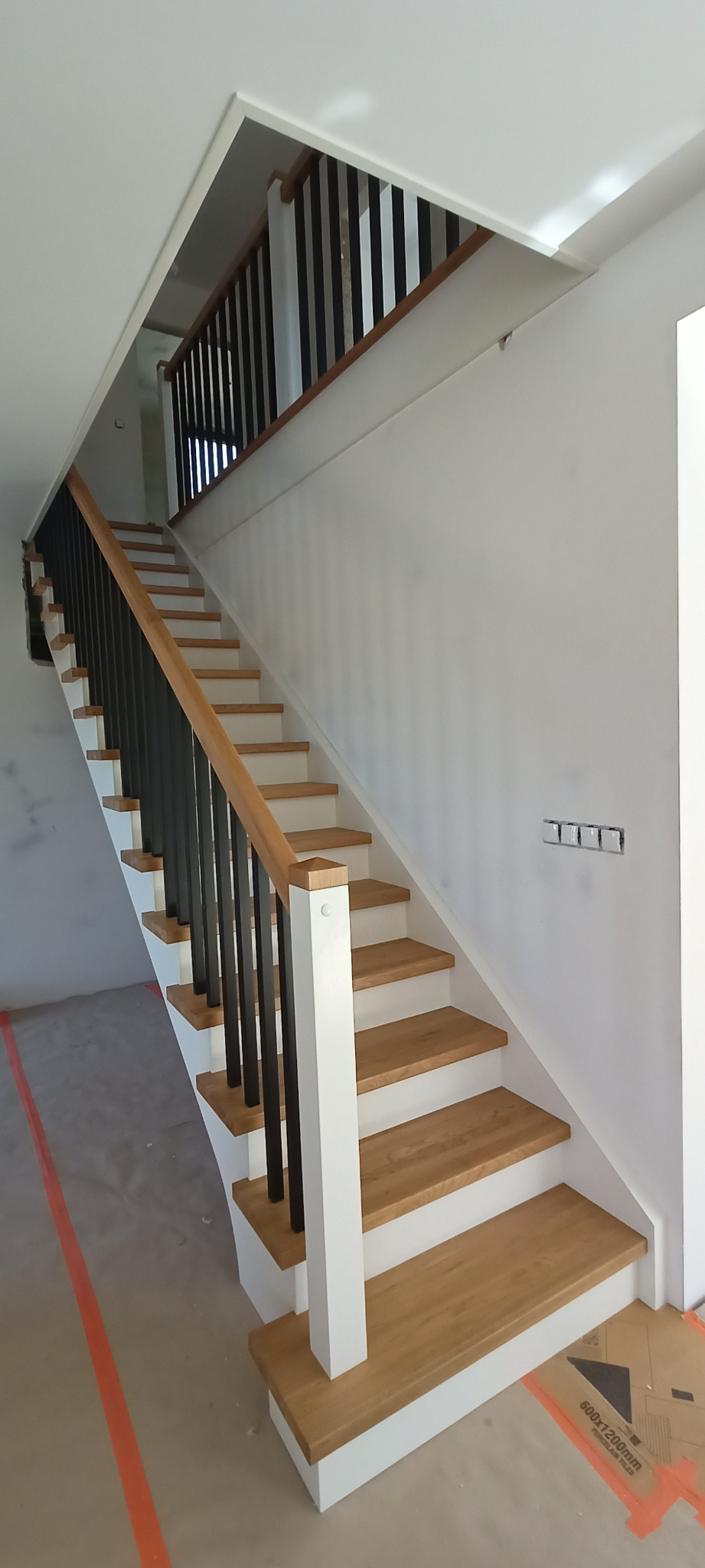 Nowoczesne schody z drewnianymi stopniami i czarną balustradą w jasnym wnętrzu. Widoczne detale wykończenia i minimalistyczny design.