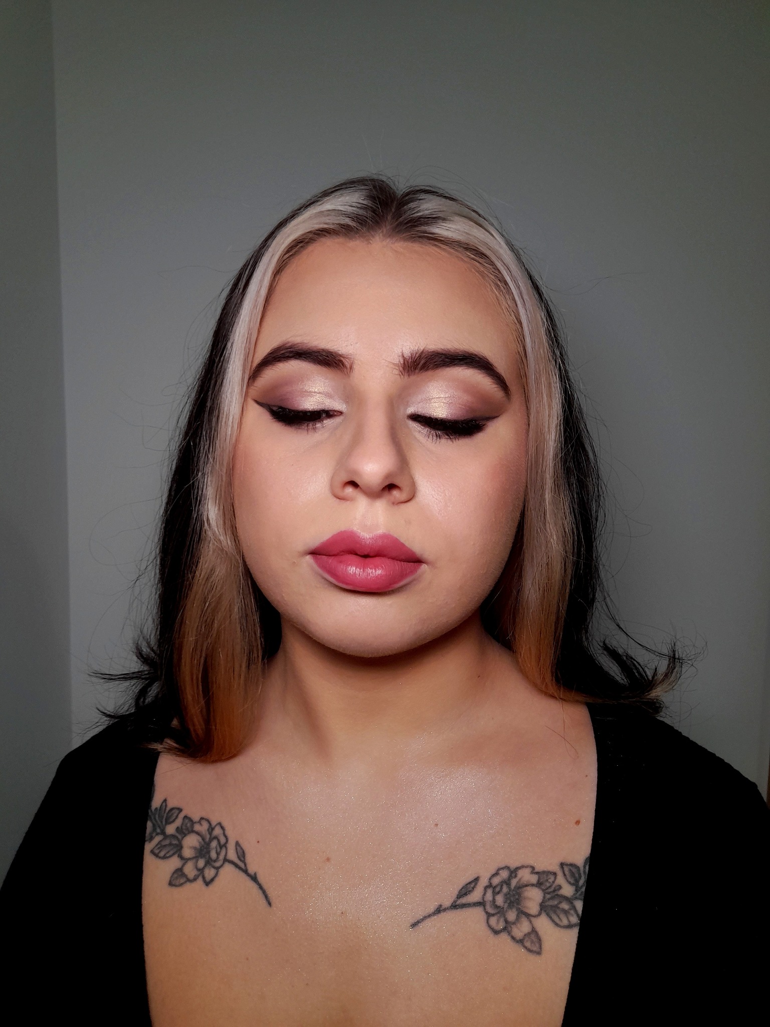 Portret kobiety z zamkniętymi oczami, prezentujący wykonany delikatny, błyszczący make-up oczu z czarną kreską oraz pomalowane na różowo usta; widoczne tatuaże kwiatowe na dekolcie oraz pasmo...