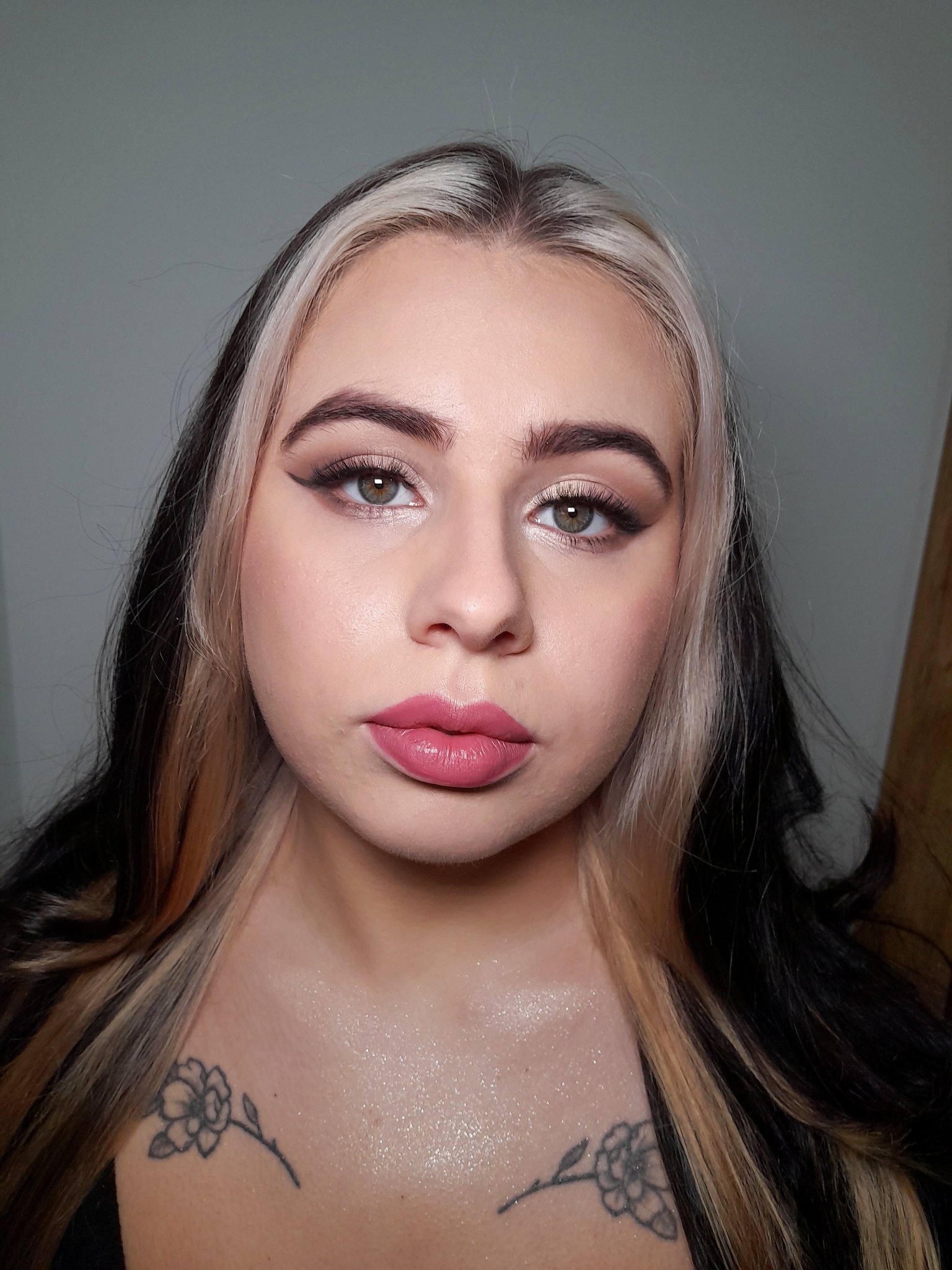 Portret kobiety z profesjonalnie wykonanym dziennym wizażem, podkreślone oczy czarnym eyelinerem i różowe usta, widoczny tatuaż kwiatowy na dekolcie, włosy z pasemkami blond i czarnymi.