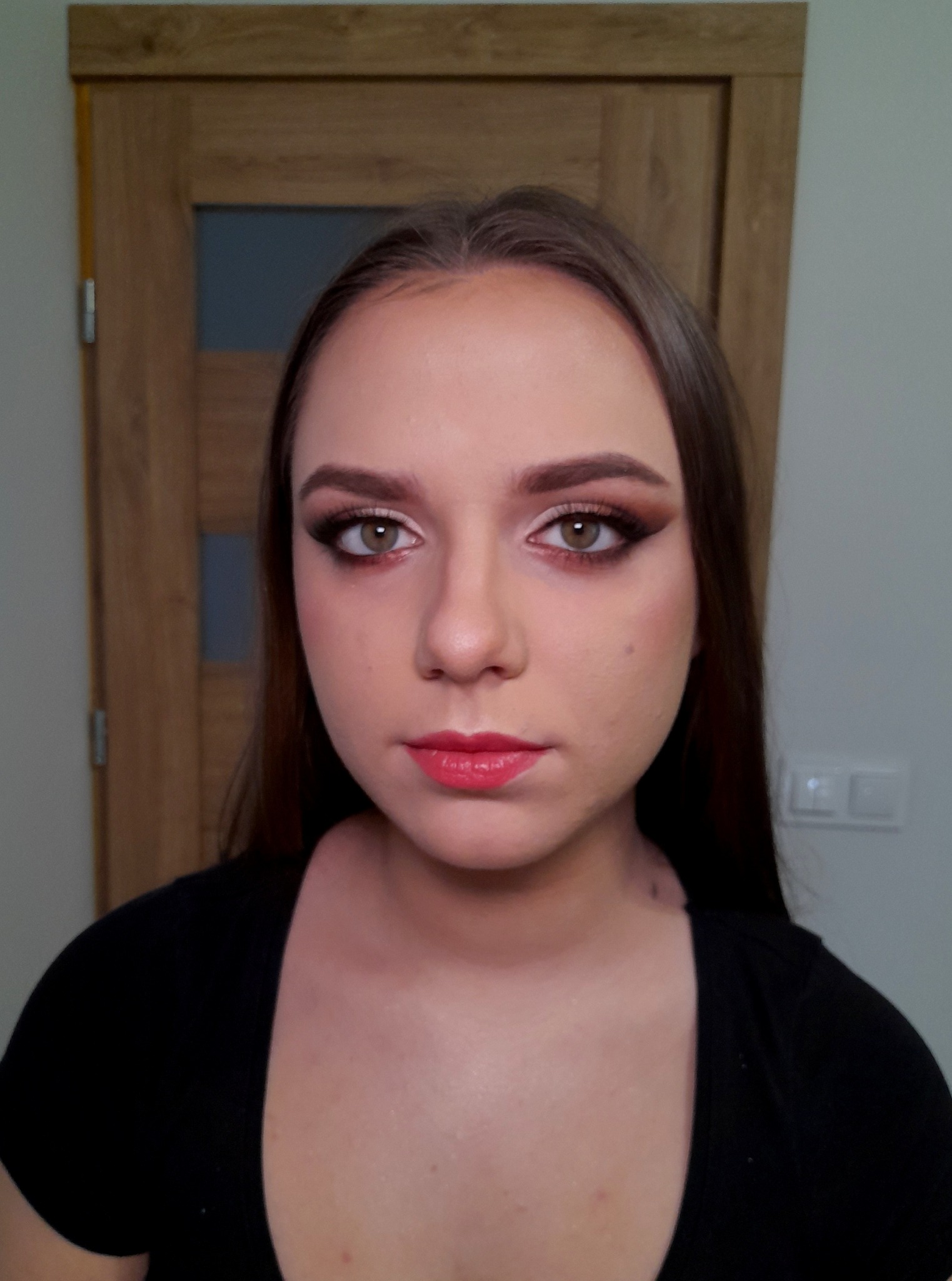 Portret młodej kobiety z brązowym cieniem do powiek, wyraźnym eyelinerem i koralową pomadką, na tle drzwi i ściany.