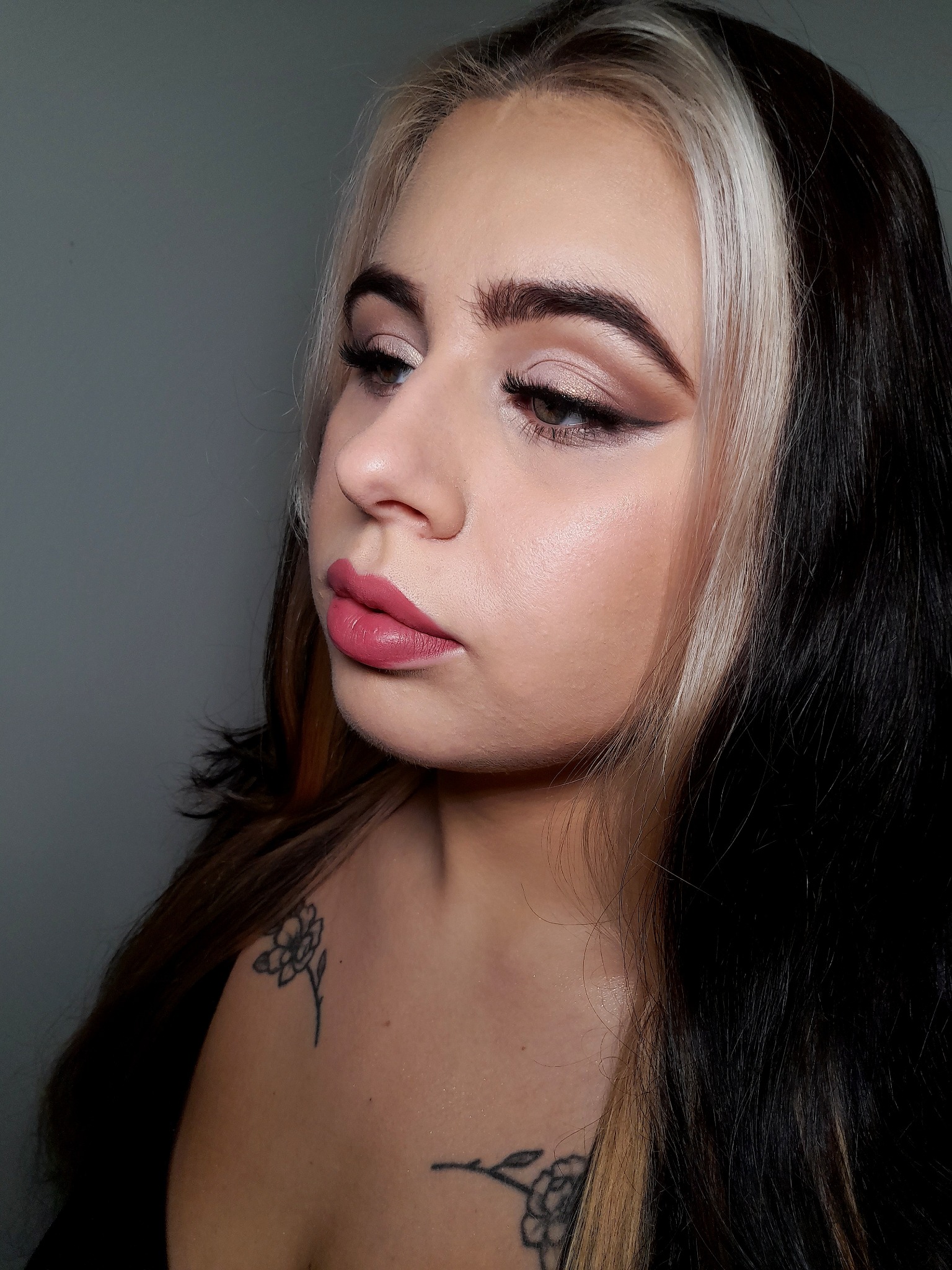 Portret kobiety z blond pasmem włosów, delikatnym makijażem oczu z eyelinerem i różowymi ustami, widoczne tatuaże kwiatowe na ramieniu i dekolcie.
