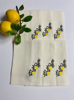 Kremowe serwetki z haftowanymi gałązkami cytryn, ułożone obok świeżych cytryn z liśćmi. Delikatny, estetyczny wzór haftu.