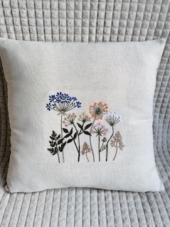Beżowa poduszka z ręcznie wykonanym, kolorowym haftem przedstawiającym łąkę z kwitnącymi roślinami na tle teksturowanej tkaniny.
