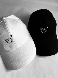 Dwie czapki z daszkiem: biała i czarna, z delikatnym wzorem serca wykonanym nićmi. Stonowana kolorystyka, minimalistyczny design. Precyzyjne wykonanie.