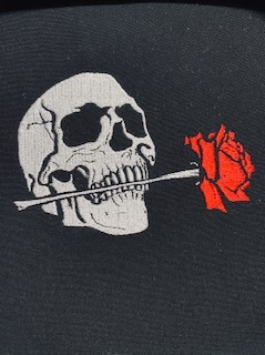 Czarna tkanina z wyhaftowanym białym czaszką trzymającą czerwoną różę w zębach. Detal haftu, widoczne ściegi.
