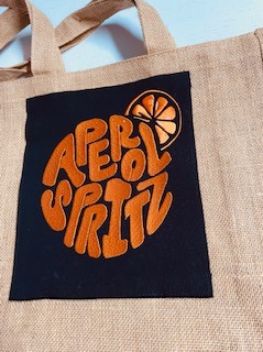Lniana torba z czarną naszywką i haftowanym logo Aperol Spritz w odcieniach pomarańczy. Widoczne detale haftu i tekstury lnu. Jasne, równomierne oświetlenie.