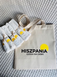 Lniana torba z haftowanym logo 'Hiszpania czeka na Ciebie' i trzy białe ręczniki z haftowanym cytrusem, leżące na szarej, pikowanej narzucie.