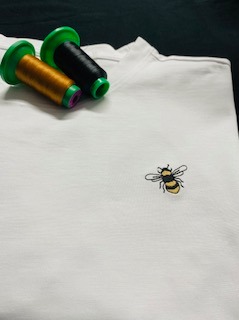 Biała koszulka z haftowaną pszczołą, obok szpulki nici w kolorze złotym i czarnym. Detal haftu i nici, bliski kadr.