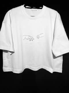 Biała koszulka z krótkim rękawem, z minimalistycznym czarnym nadrukiem przedstawiającym dłonie, wisząca na czarnym wieszaku na tle ciemnej ściany.