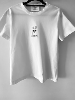 Biała koszulka z czarnym, minimalistycznym wzorem lamy w okularach, zawieszona na wieszaku. Stonowana kolorystyka. Precyzyjne wykonanie wzoru.