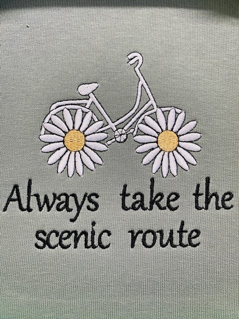 Jasnozielona tkanina z dekoracyjnym, maszynowym motywem roweru z kwiatów i napisem 'Always take the scenic route' wykonanym czarną nicią.