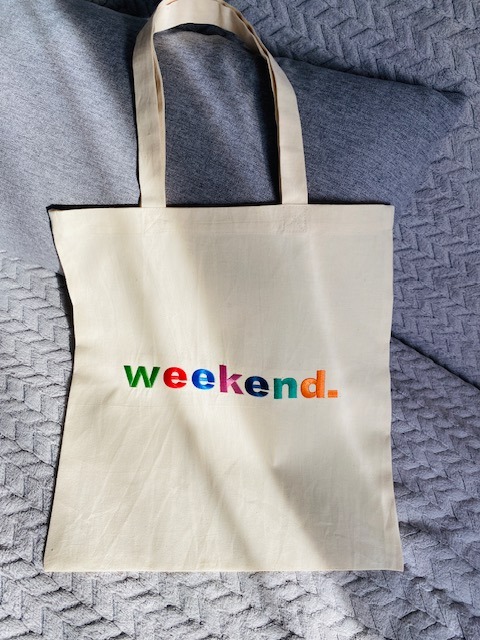 Beżowa torba z tkaniny z kolorowym napisem 'weekend.' leży na szarej teksturowanej powierzchni. Widoczne uchwyty torby i delikatne załamania materiału.