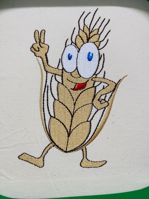 Uśmiechnięty, haftowany kłos zboża z oczami i rękami, gestykulujący na jasnym tle. Detalowe szwy i żywe kolory. Zabawny motyw.