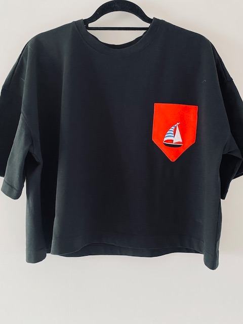 Czarna bluzka z krótkim rękawem i czerwoną kieszenią z motywem żaglówki, wisząca na czarnym wieszaku na tle białej ściany.