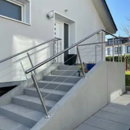 Zewnętrzne schody z szarego kamienia z nowoczesną balustradą ze stali nierdzewnej z cienkimi linkami, prowadzące do białych drzwi wejściowych domu.