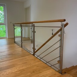 GSsteel Jarosław Sordyl - Nowoczesna balustrada ze szkła i stali nierdzewnej z drewnianymi poręczami, widok z góry na drewnianą podłogę i fragment schodów, jasne wnętrze z oknem.