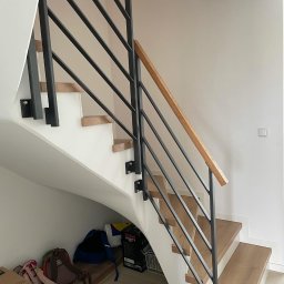 GSsteel Jarosław Sordyl - Nowoczesne schody z drewnianymi stopniami i czarną metalową balustradą z drewnianą poręczą, widok z dołu na tle białej ściany, pod schodami widoczne przedmioty.