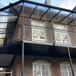 GSsteel Jarosław Sordyl - Zewnętrzny balkon z czarną metalową konstrukcją, balustradą z ozdobnymi elementami i przezroczystym zadaszeniem, zamontowany na ceglanym budynku z oknami zabezpieczonymi folią.