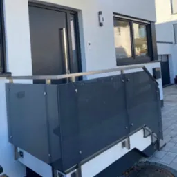 Nowoczesny balkon z szarą szklaną balustradą i nierdzewną poręczą, zamontowaną na białej elewacji budynku.