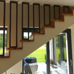 GSsteel Jarosław Sordyl - Nowoczesne schody z drewnianymi stopniami i minimalistyczną, czarną metalową balustradą z prostokątnymi elementami pionowymi, widok z dołu na otwarte drzwi tarasowe i ogród.