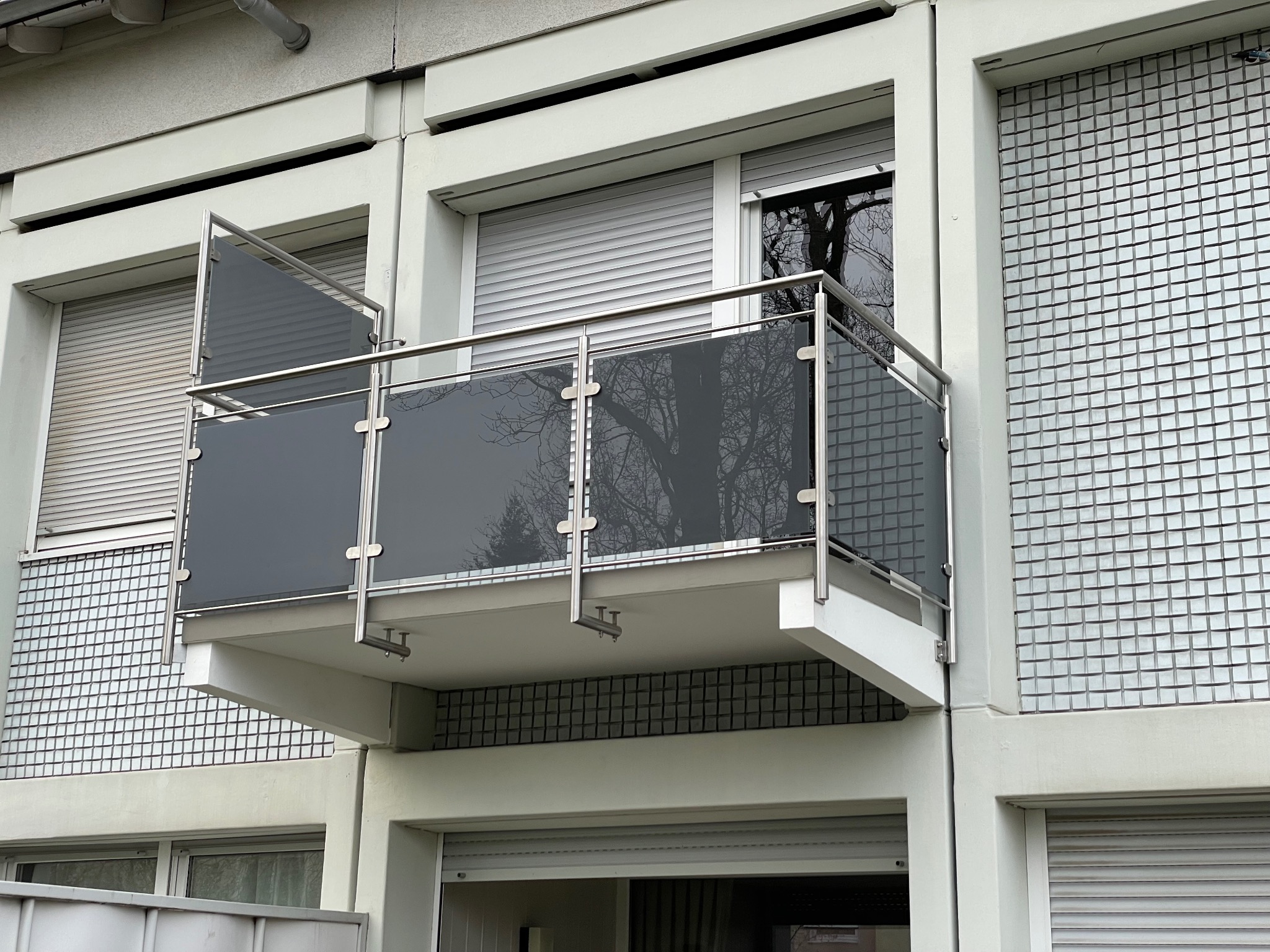 Balkon z szarymi szklanymi panelami balustrady i nierdzewnymi elementami mocującymi, widok z dołu na tle budynku z roletami.