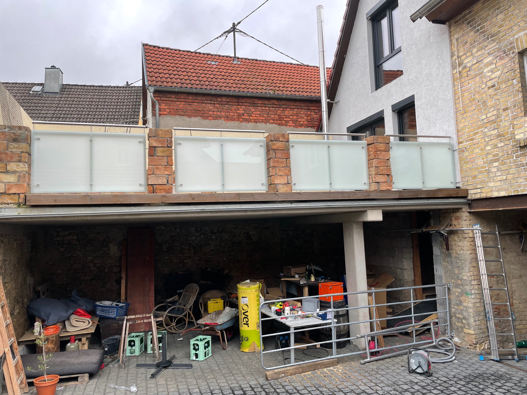 Balkon z balustradą ze szkła matowego i nierdzewnej stali, wsparty na ceglanych słupkach, widok z dołu na otwarty garaż z narzędziami i materiałami budowlanymi.
