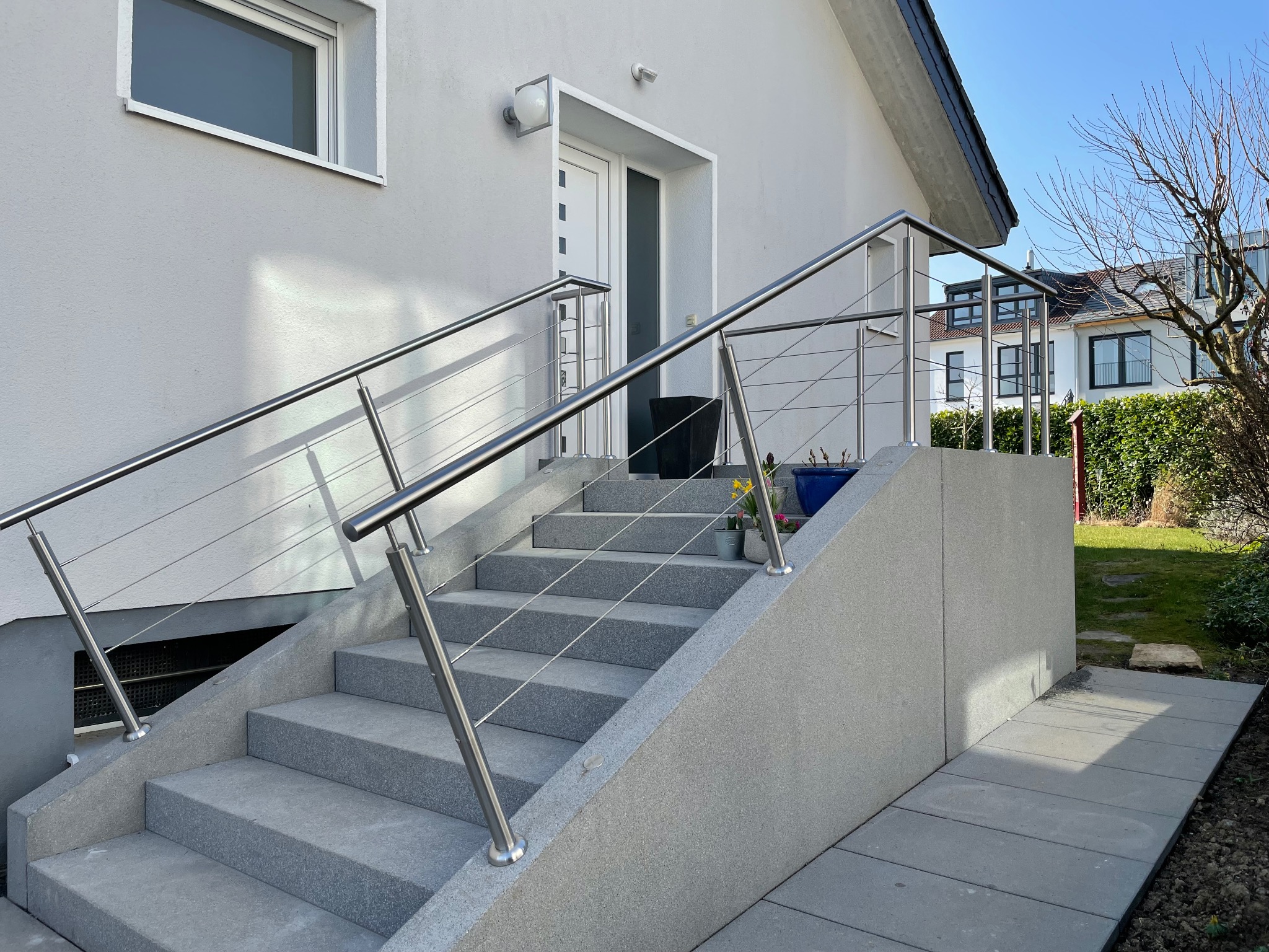 Zewnętrzne schody z szarego kamienia prowadzące do domu, zabezpieczone nowoczesną balustradą ze stali nierdzewnej z linkami, z widoczną roślinnością w tle.