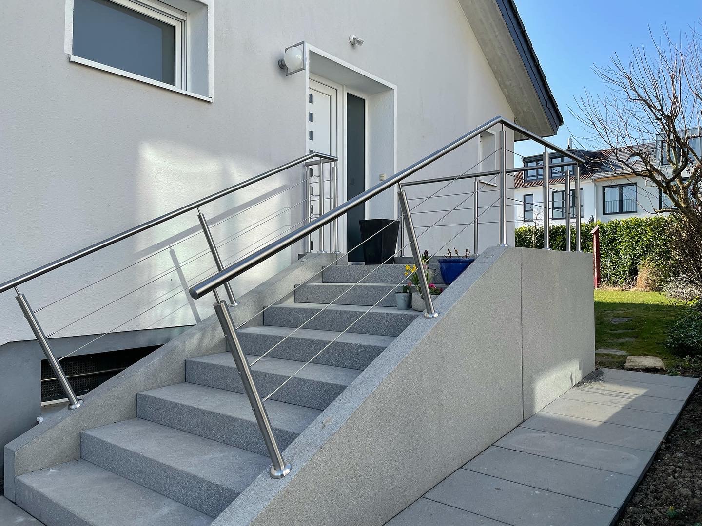 Zewnętrzne schody z szarego kamienia z nowoczesną balustradą ze stali nierdzewnej z cienkimi linkami, prowadzące do białych drzwi wejściowych domu.