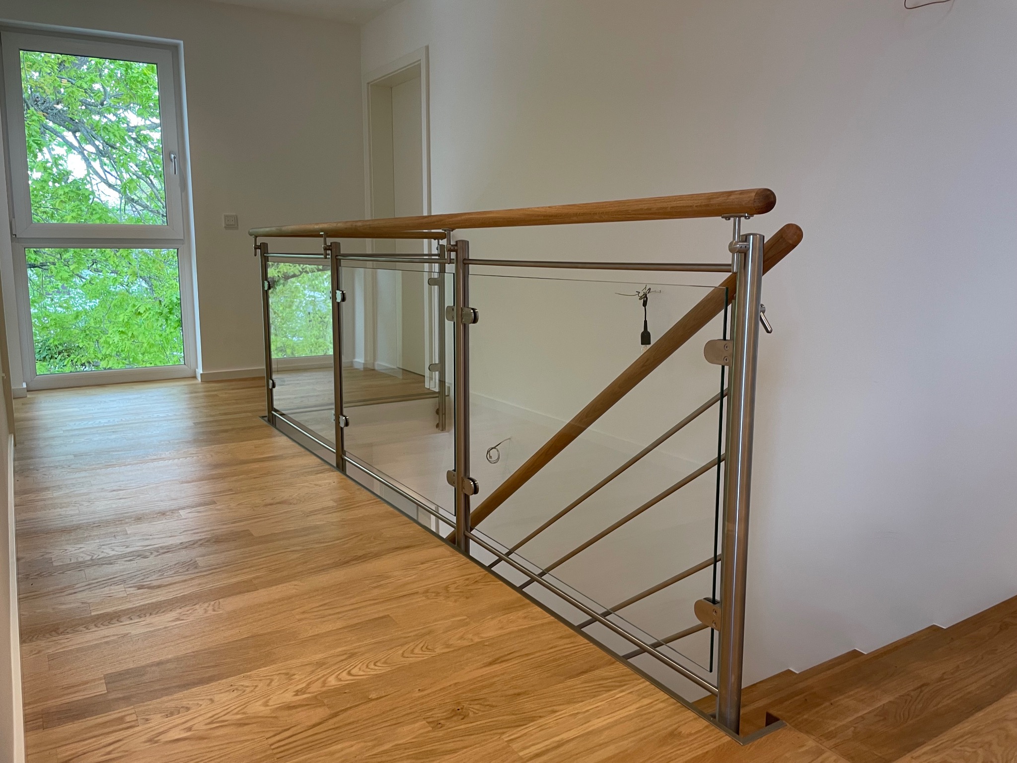 Nowoczesna balustrada ze szkła i stali nierdzewnej z drewnianymi poręczami, widok z góry na drewnianą podłogę i fragment schodów, jasne wnętrze z oknem.