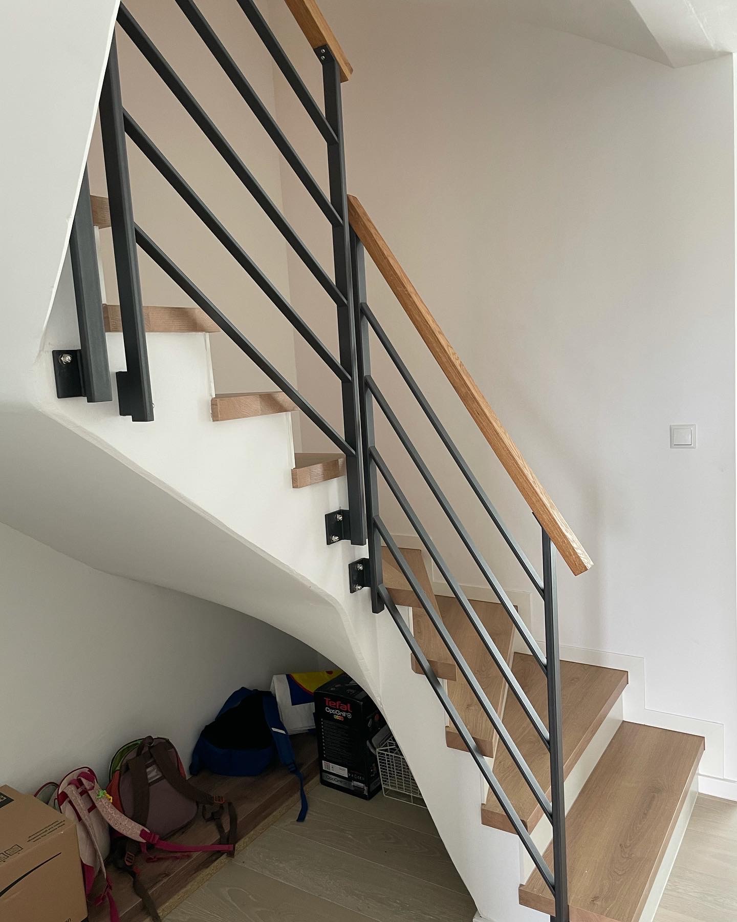 Nowoczesne schody z drewnianymi stopniami i czarną metalową balustradą z drewnianą poręczą, widok z dołu na tle białej ściany, pod schodami widoczne przedmioty.