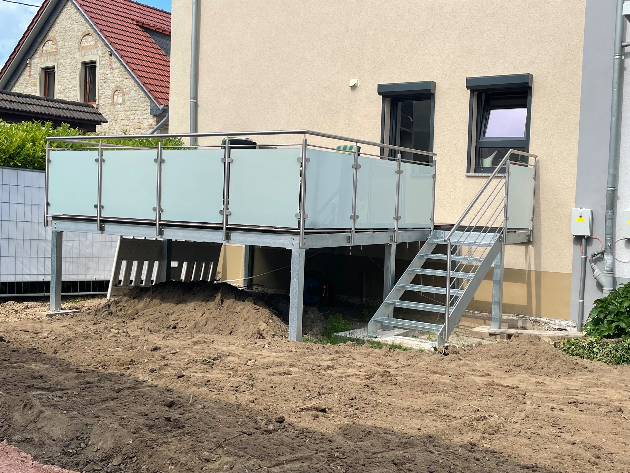 Zewnętrzny balkon z metalową konstrukcją, schodami i mlecznymi szklanymi balustradami, widok z poziomu gruntu na tle budynku mieszkalnego i przygotowanego terenu.