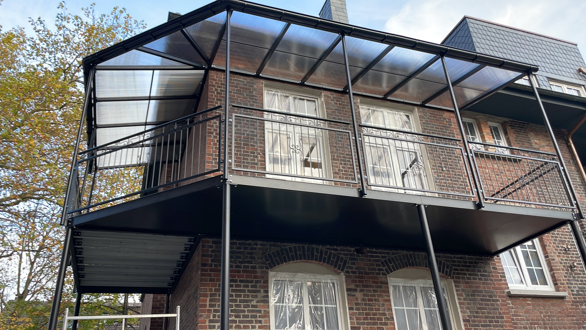 Zewnętrzny balkon z czarną metalową konstrukcją, balustradą z ozdobnymi elementami i przezroczystym zadaszeniem, zamontowany na ceglanym budynku z oknami zabezpieczonymi folią.