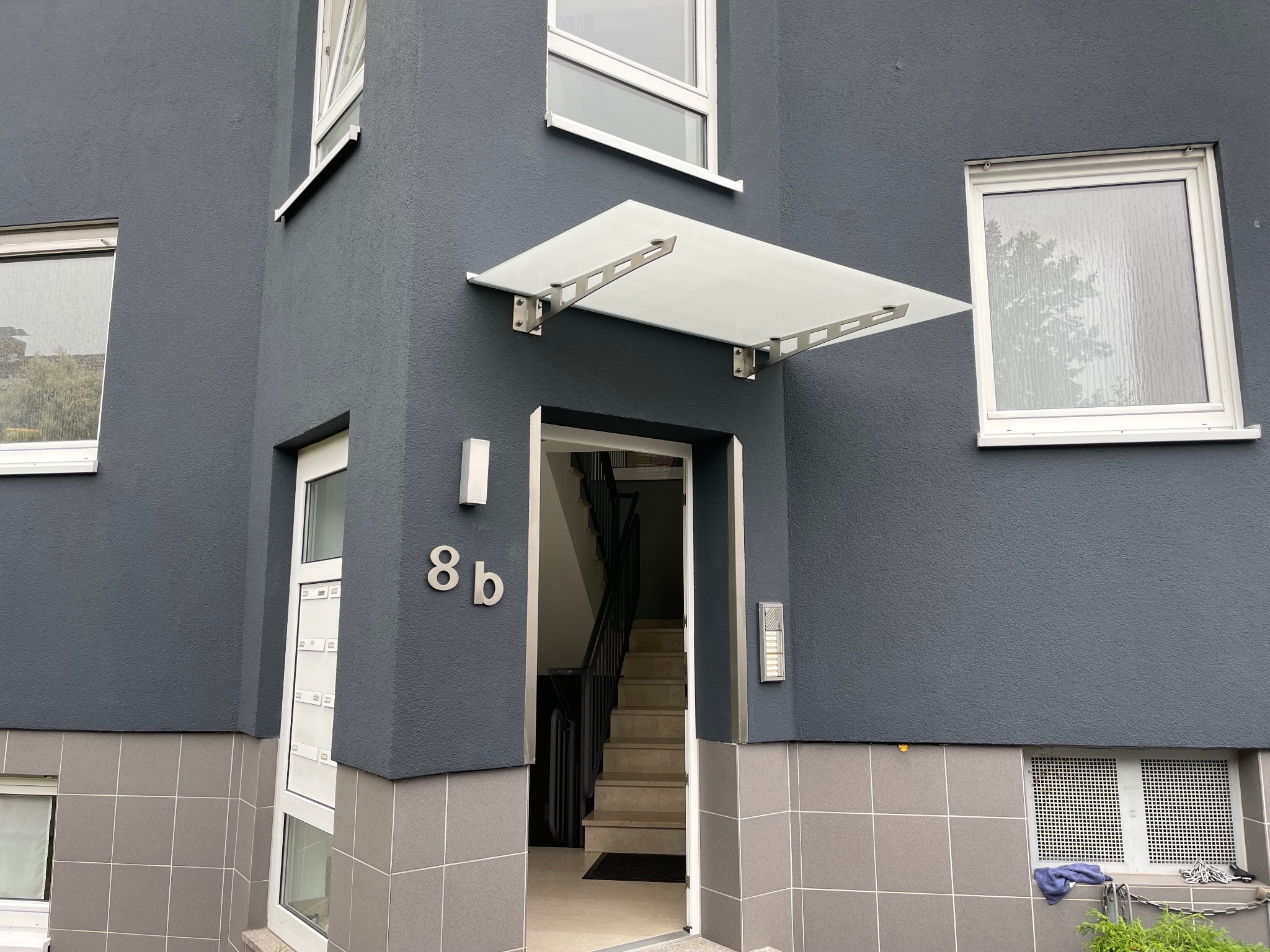 Wejście do budynku z szarą elewacją, osłonięte szklanym daszkiem na metalowej konstrukcji, widoczne schody wewnątrz.