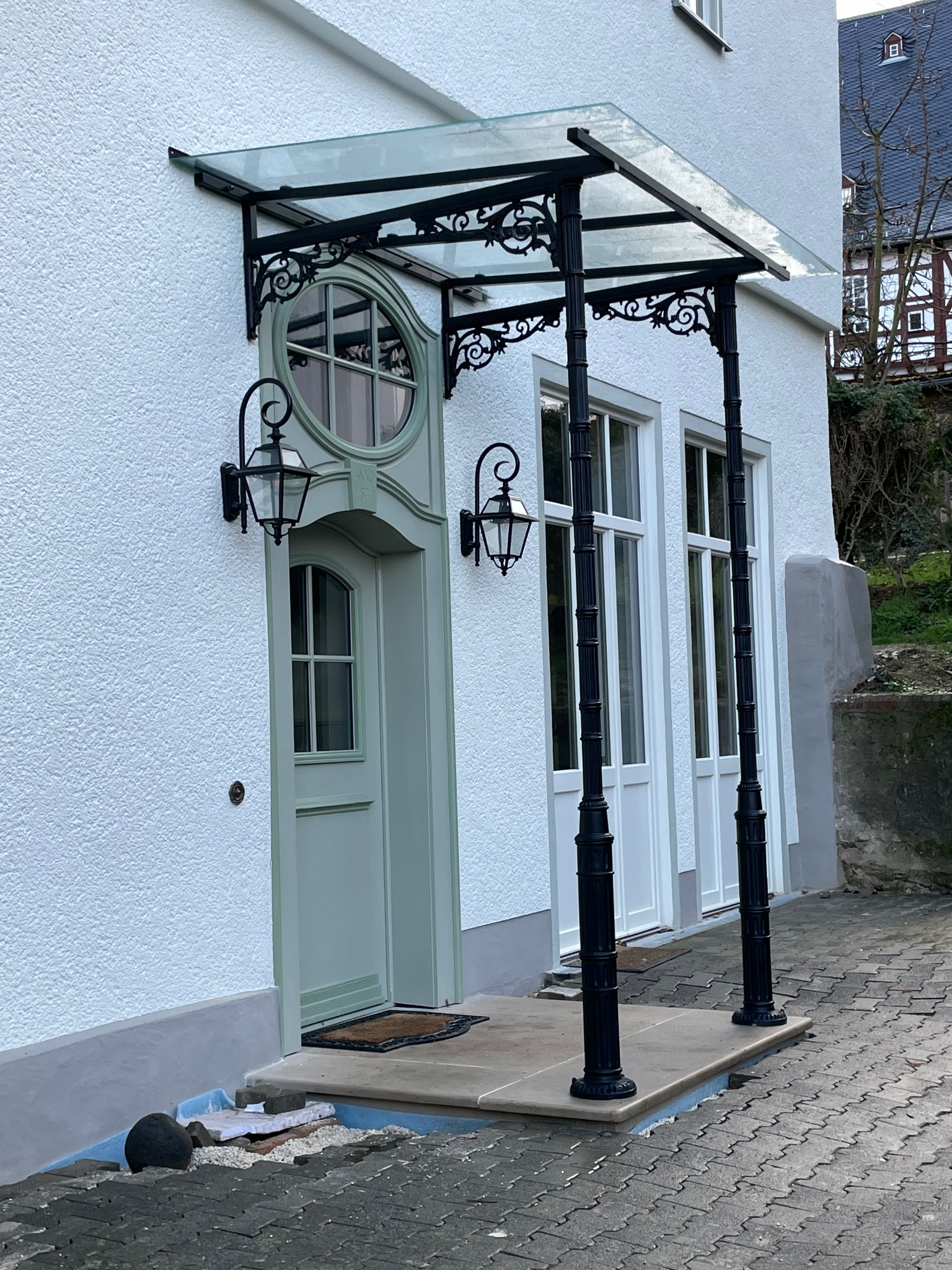 Eleganckie zadaszenie nad wejściem do domu z ozdobną, metalową konstrukcją i szklanym dachem, wsparte na kolumnach, z widocznymi detalami dekoracyjnymi i lampami ściennymi.