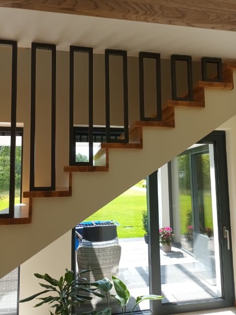 Nowoczesne schody z drewnianymi stopniami i minimalistyczną, czarną metalową balustradą z prostokątnymi elementami pionowymi, widok z dołu na otwarte drzwi tarasowe i ogród.