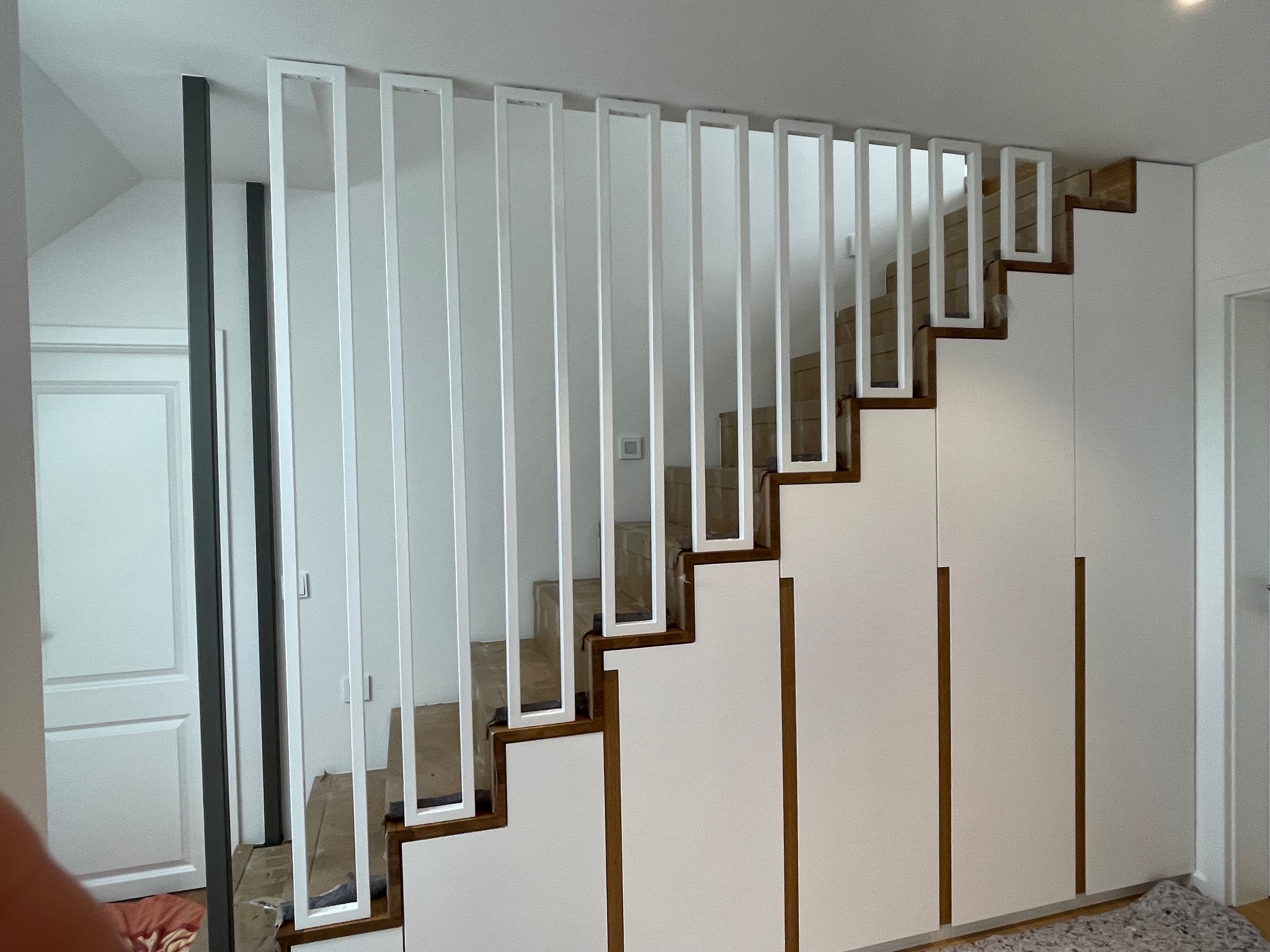 Nowoczesne schody drewniane z białymi, pionowymi elementami balustrady, zintegrowane z białą zabudową szaf wnękowych.