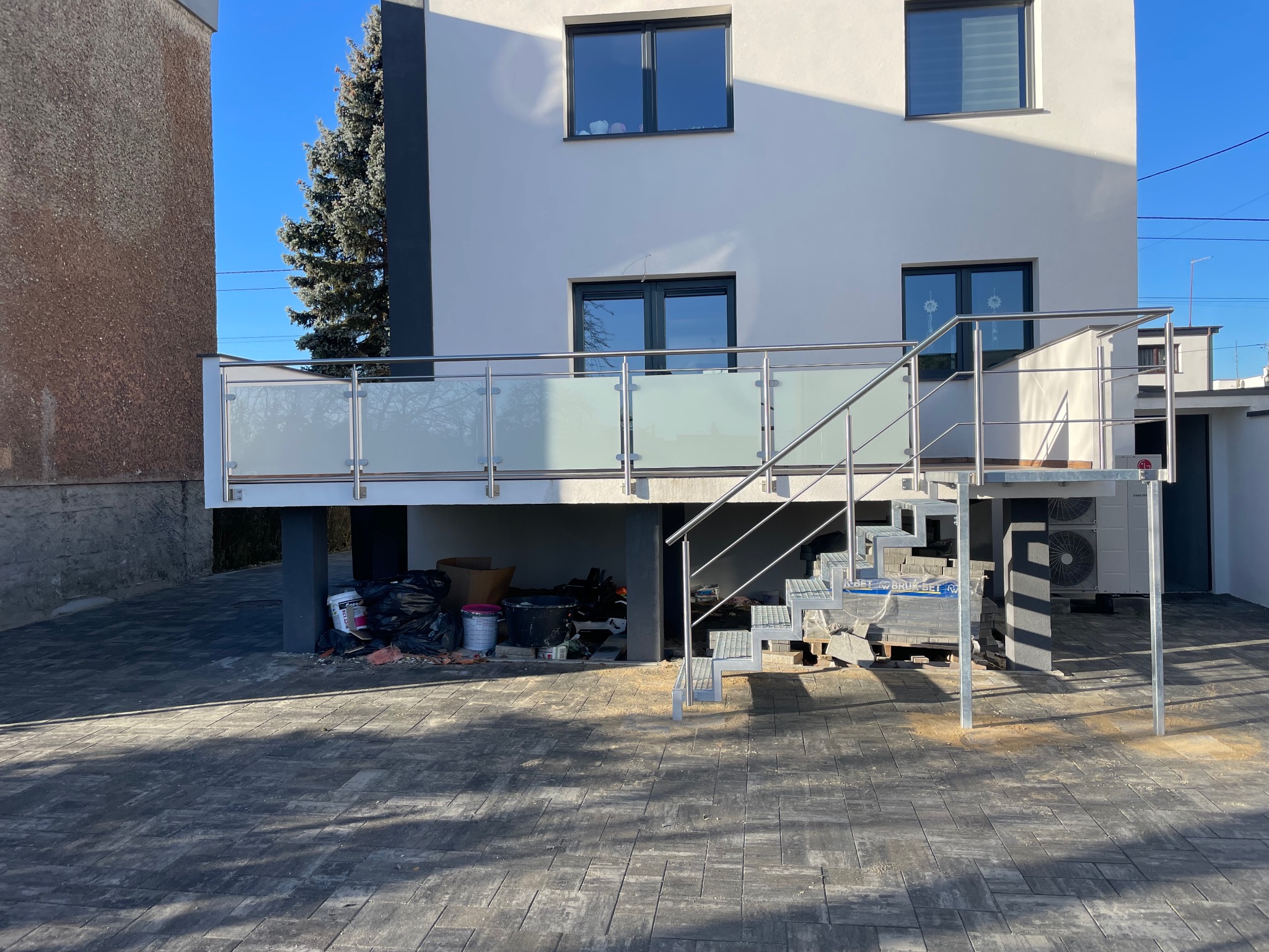 Zewnętrzne schody metalowe z ażurowymi stopniami prowadzące na balkon z balustradą szklaną, widoczne materiały budowlane i elementy instalacji grzewczej.